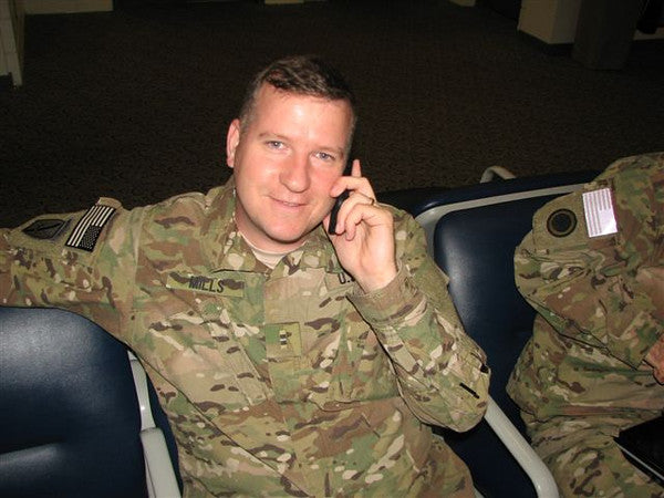 Arthur Mills in military uniform, seated in an airport terminal area, smiling while talking on a cell phone. An American flag patch is visible on his sleeve.